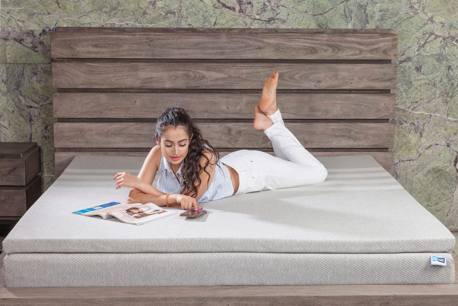 A young woman relaxing on a bed with a magazine and cellphone, set against a stylish wooden headboard.