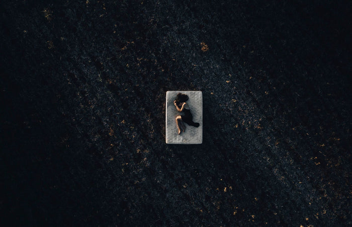 Aerial view of a woman in a black dress lying peacefully on a mattress in a dark open field at night.