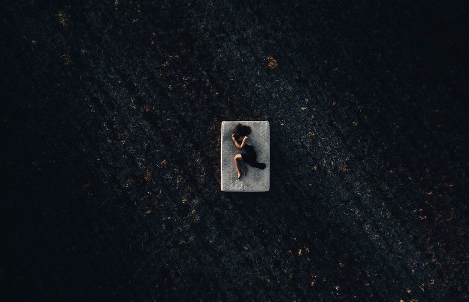 Aerial view of a woman in a black dress lying peacefully on a mattress in a dark open field at night.
