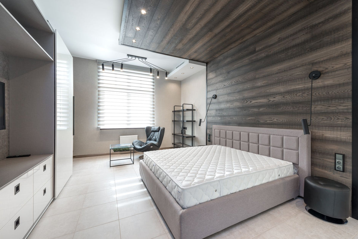 Bed with white mattress placed at wall with decorative element against cupboards in light stylish bedroom with window and armchair
