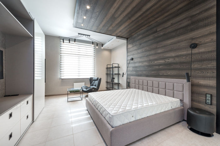 Bed with white mattress placed at wall with decorative element against cupboards in light stylish bedroom with window and armchair