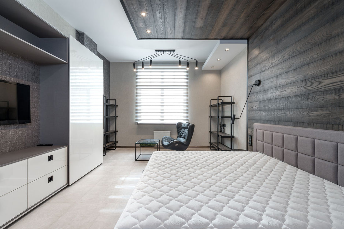 Bed with mattress placed against wall with modern TV and white cupboards in stylish bedroom with window and leather armchair