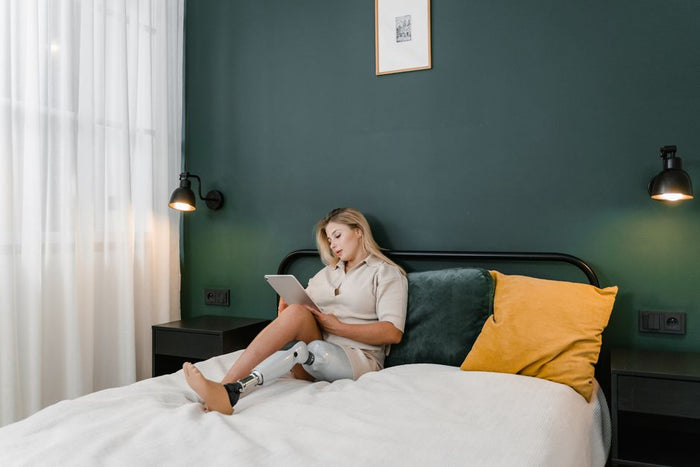 A woman with a prosthetic leg sits on a bed in a modern bedroom, using a tablet.