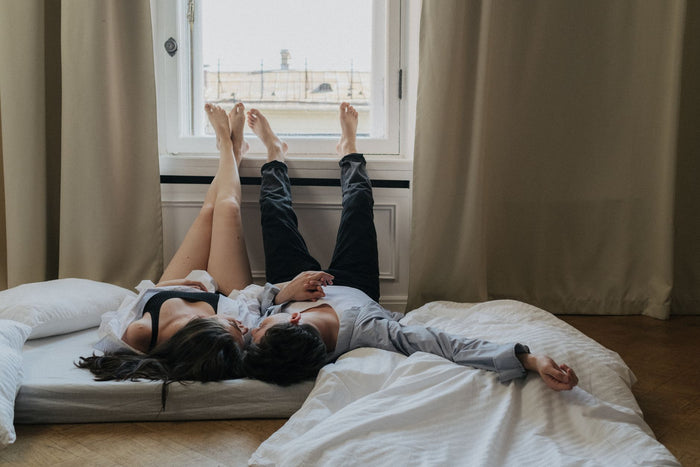 A romantic couple lies on a mattress with legs raised by the window, enjoying cozy intimate moments.
